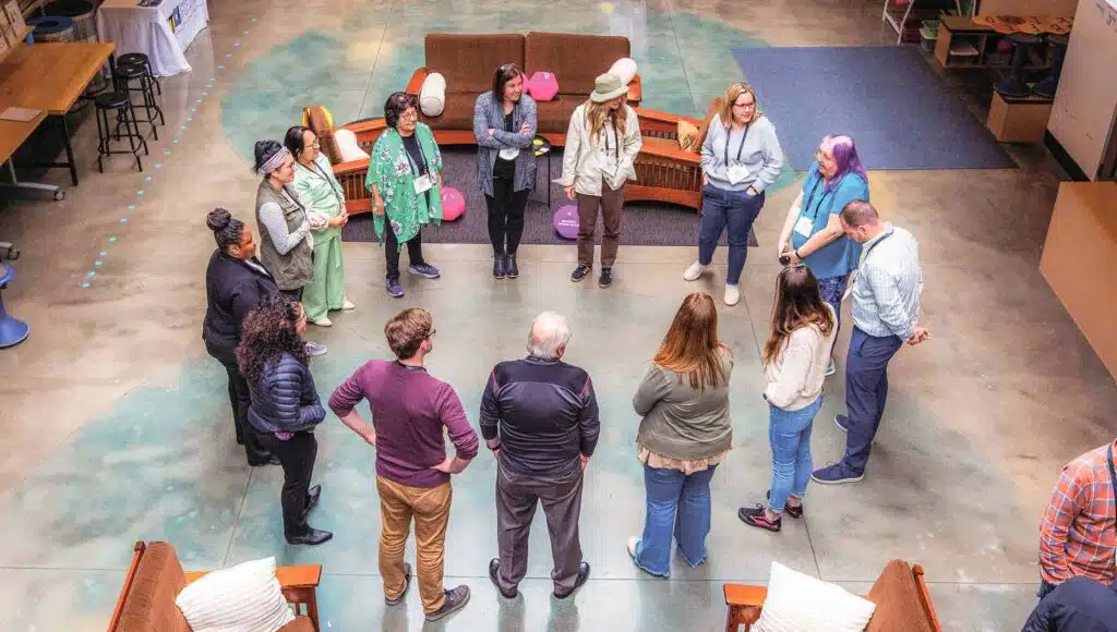 A group of people stands in a circle having a discussion during the in-person PBL Design Camp in a spacious indoor area, with sofas and tables visible around them. The photo is taken from above.