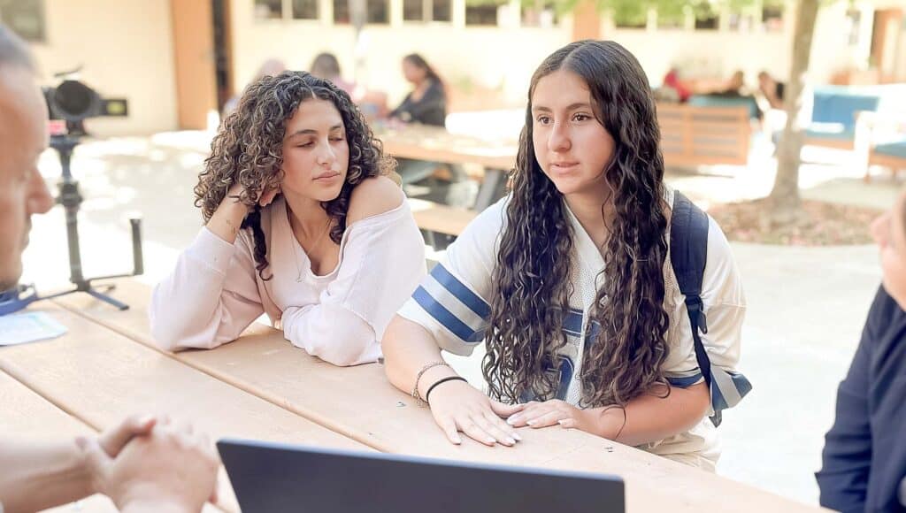Two young women sit at an outdoor table, engaged in conversation with two people off-camera. One woman is speaking while the other listens attentively. A laptop and a camera are visible on the table.
