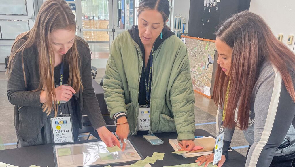 Three women wearing visitor badges stand around a table, examining and organizing paper cards and documents. One woman points at a card while the others watch and discuss. They appear to be collaborating on a project indoors.