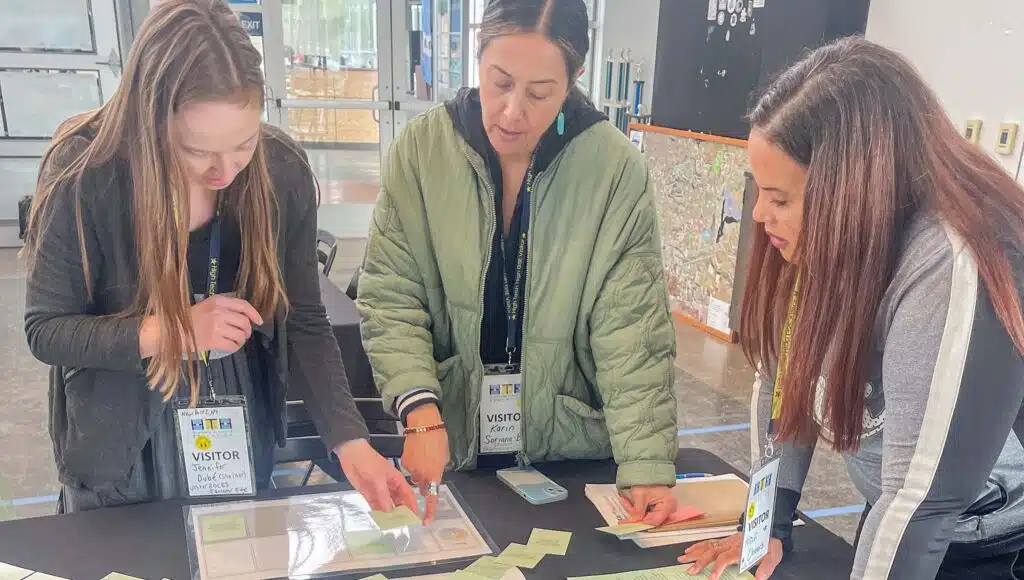 Three women wearing visitor badges stand around a table, examining and organizing paper cards and documents. One woman points at a card while the others watch and discuss. They appear to be collaborating on a project indoors.