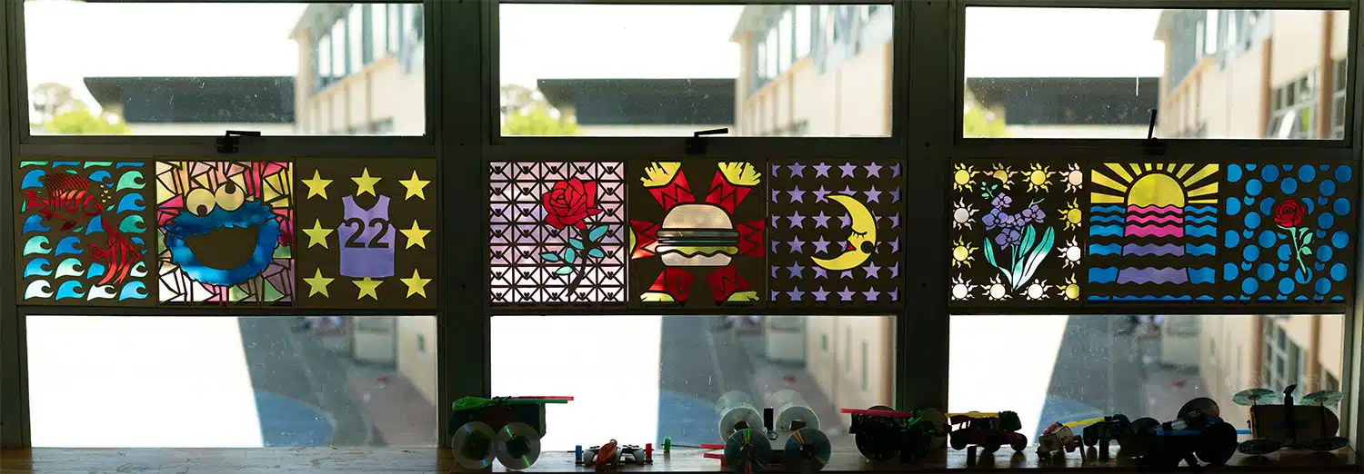 Colorful paper art panels featuring stars, flowers, a rose, a burger, a crescent moon, and a sunrise create a vibrant student work display on the window. Small toy vehicles line the windowsill below with buildings visible outside.