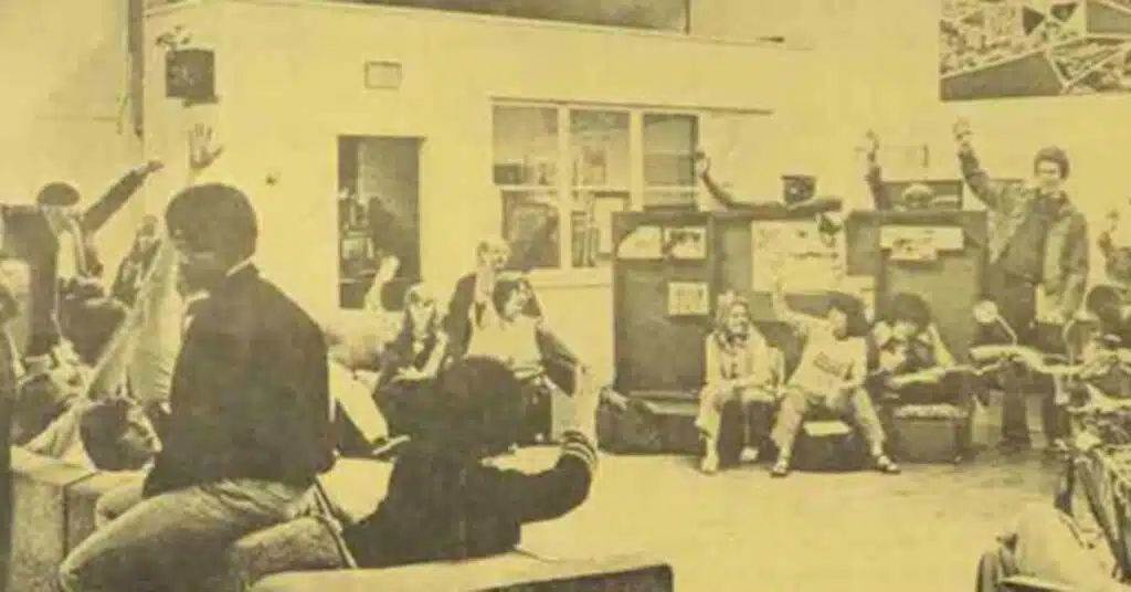 A group of people sit and stand in a room, many with hands raised. The image has a yellowish, vintage tone, suggesting it is from a previous decade—a likely scene from an alternative high school meeting or group school discussion.