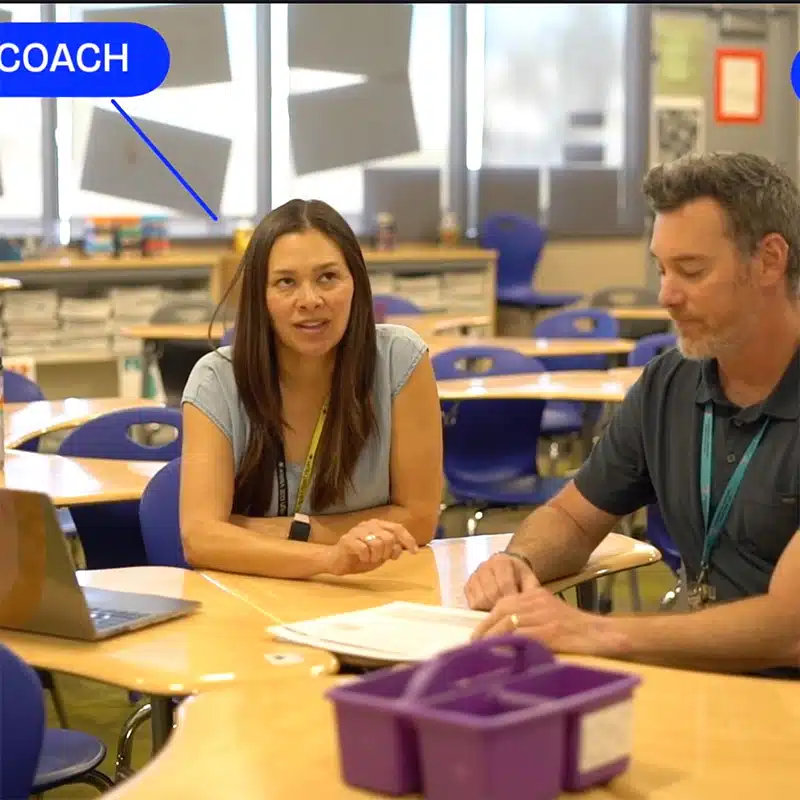 A woman labeled COACH sits at a classroom table discussing Student Work Protocol with a man. Both have papers in front of them. The classroom is empty, with blue chairs and sunlight streaming through the windows.