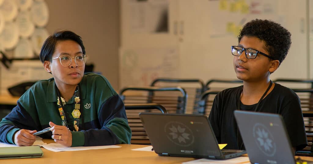 Two students sit at a table with laptops, focusing on their work. One, in a green jacket, holds a pen; the other looks thoughtful. Papers and notebooks are scattered across the table, reflecting active student involvement in learning.