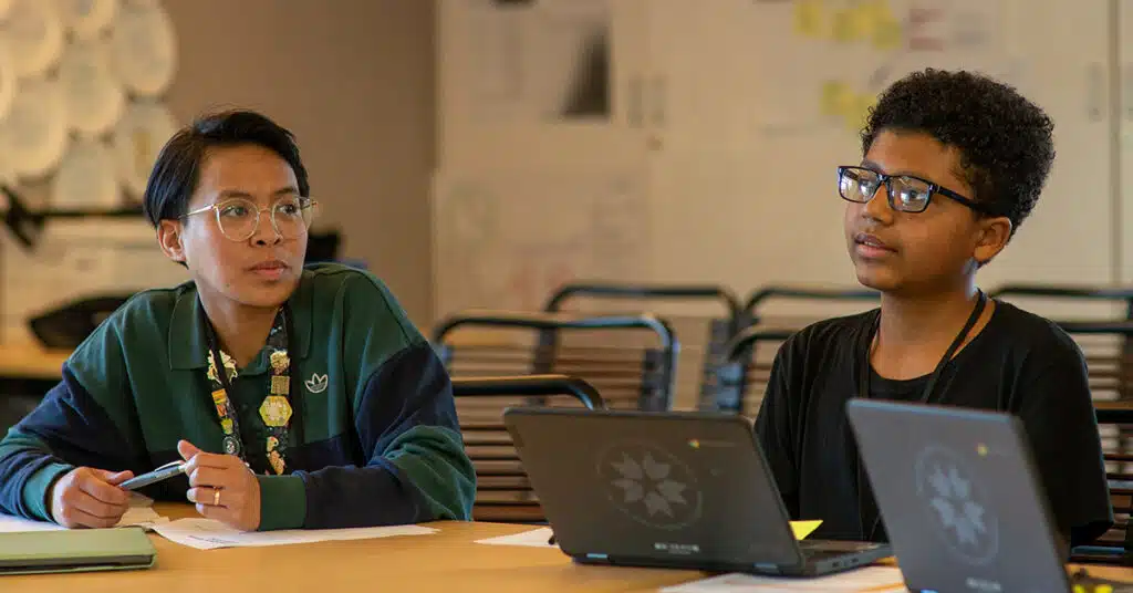 Two students sit at a table with laptops, focusing on their work. One, in a green jacket, holds a pen; the other looks thoughtful. Papers and notebooks are scattered across the table, reflecting active student involvement in learning.