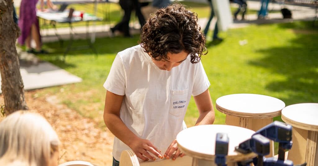 A person with curly hair, wearing a white t-shirt, focuses on working with wooden pieces outdoors on a sunny day—showcasing tangible results. Tables, trees, and blurred people are visible in the background.