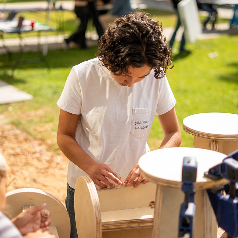 A person with curly hair wearing a white t-shirt works on assembling a wooden object outdoors on a sunny day, surrounded by round wooden pieces and tools, focused on creating tangible results in a grassy area.