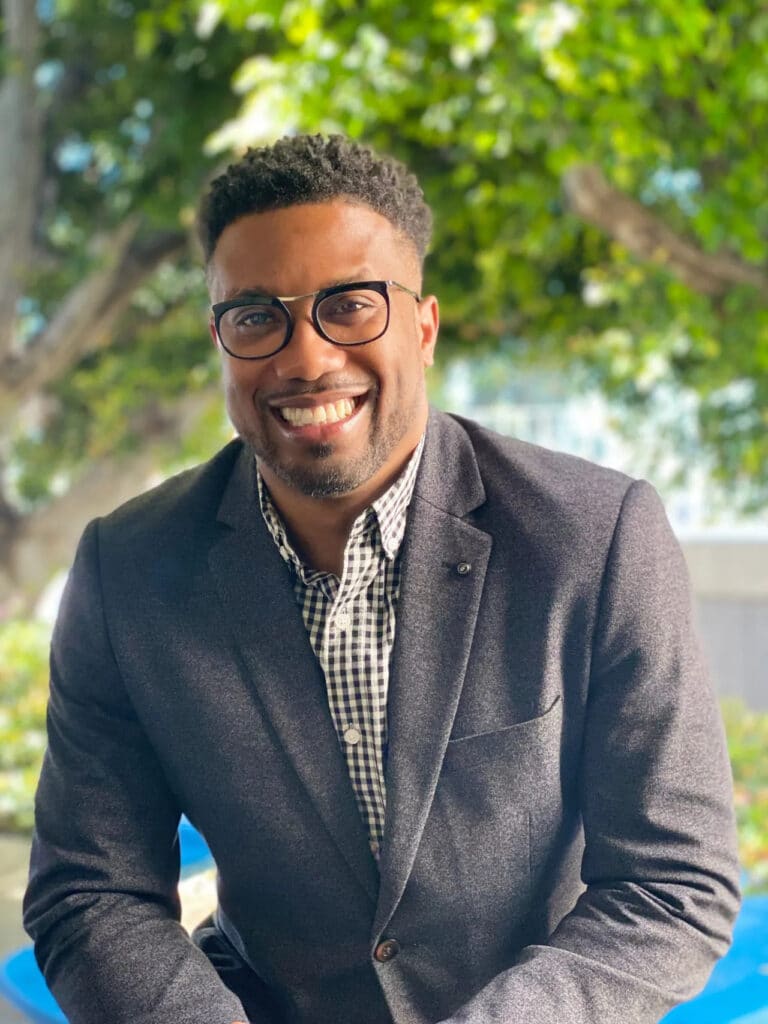A smiling man with short hair and glasses, wearing a dark blazer over a checked shirt, sits outdoors in front of green trees on a sunny day, embodying the spirit of college access and connection within the college network.
