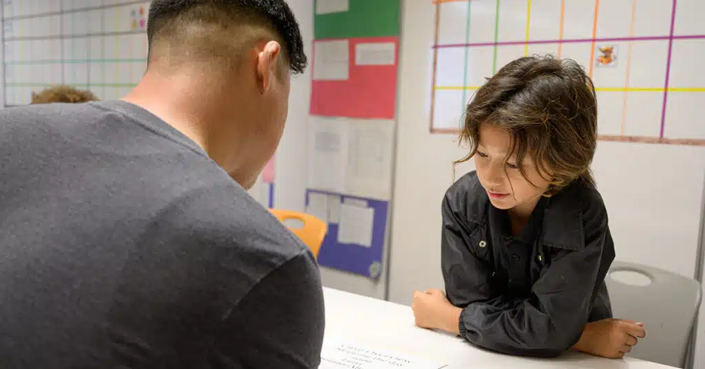An adult and a child sit at a table in a classroom, with the child looking at a paper. Colorful charts and papers focused on Learning Targets are visible on the walls behind them, creating a powerful environment for education.
