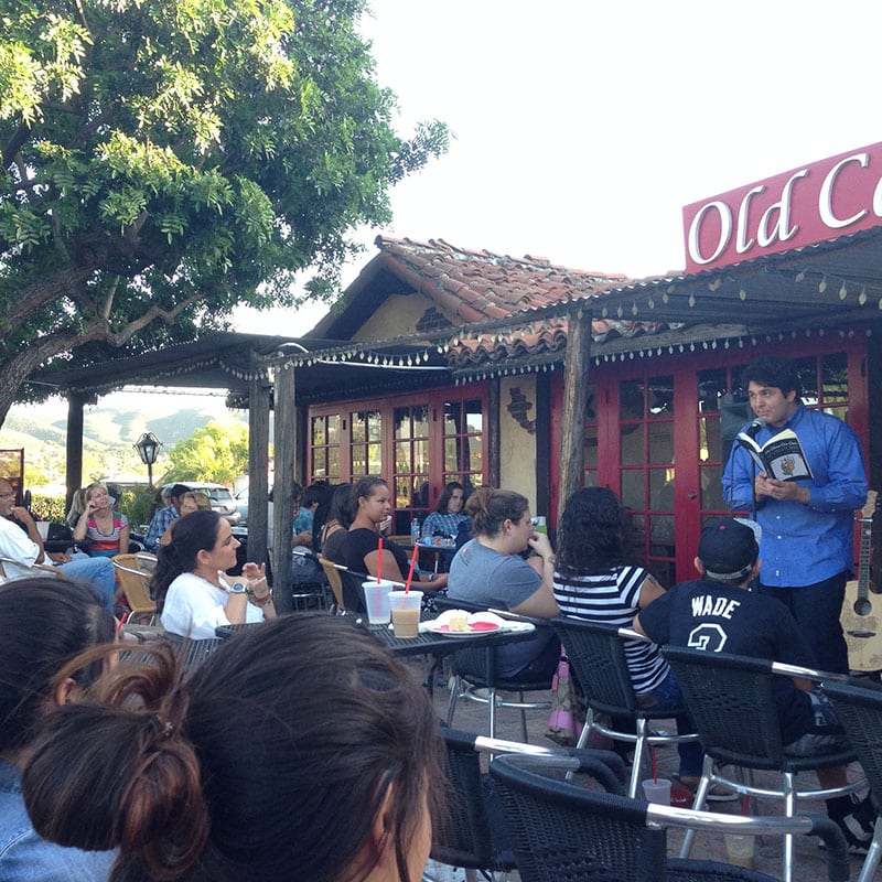A man reads from a book to an outdoor audience seated at tables outside a rustic café with red windows, under a large tree on a sunny day, turning the gathering into a simple way to connect over stories much like sharing a creative project.