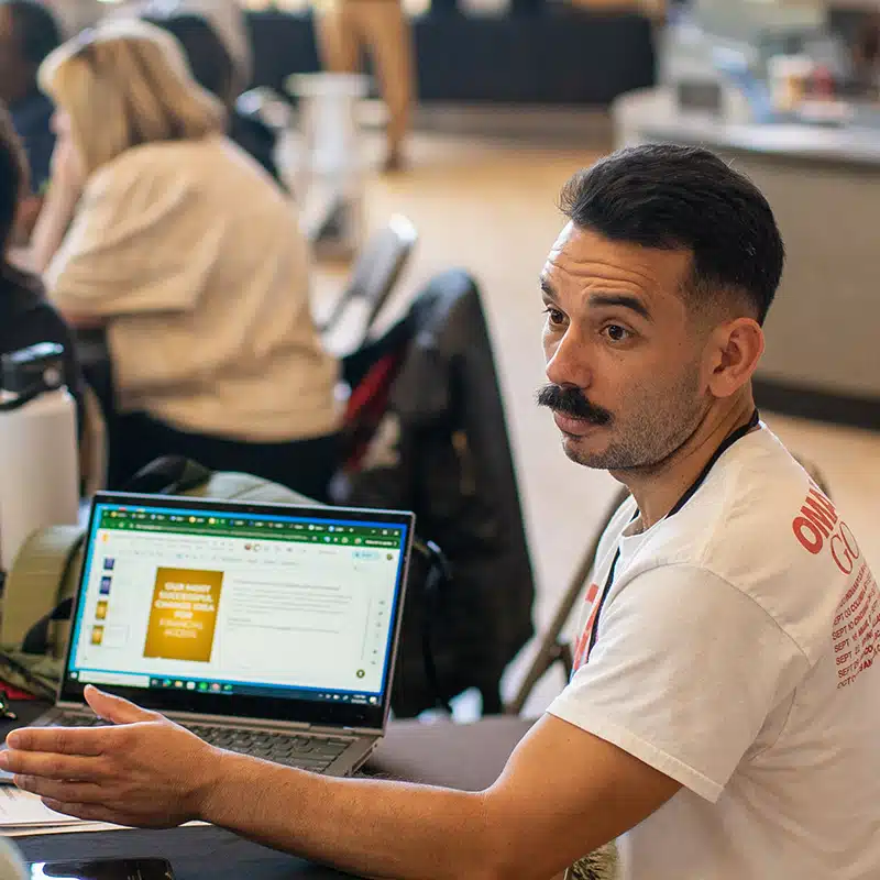 A man with a mustache sits at a table, gesturing with his hand. He faces a laptop displaying a presentation on model critiques. Other people are visible in the background, suggesting a group setting or workshop focused on continuous improvement.