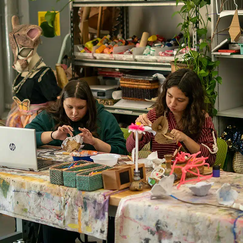 Two people work on craft projects at a table covered with art supplies, engaging in effective PBL (project-based learning) activities. Shelves of materials and a person in a rabbit costume are visible in the background. The scene appears to be an art studio or classroom.