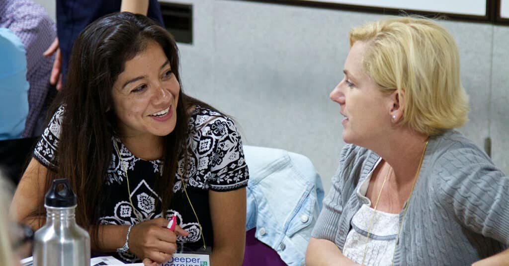 Two women sit at a table, engaged in conversation about mejora and desarrollo personal. One has dark hair and wears a patterned shirt, the other has blonde hair and wears a light cardigan. Papers and a water bottle are on the table, suggesting a thoughtful discussion focused on crecimiento and self-improvement.