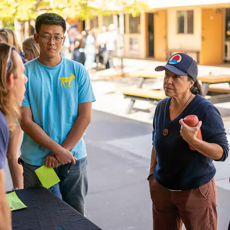 Two people stand outdoors at a table, discussing shuffling methods with someone off-camera. One holds papers, and the other holds a pink water bottle. There are benches and trees in the background, hinting at preparations to shuffle a group or discuss group segregation for an upcoming activity.