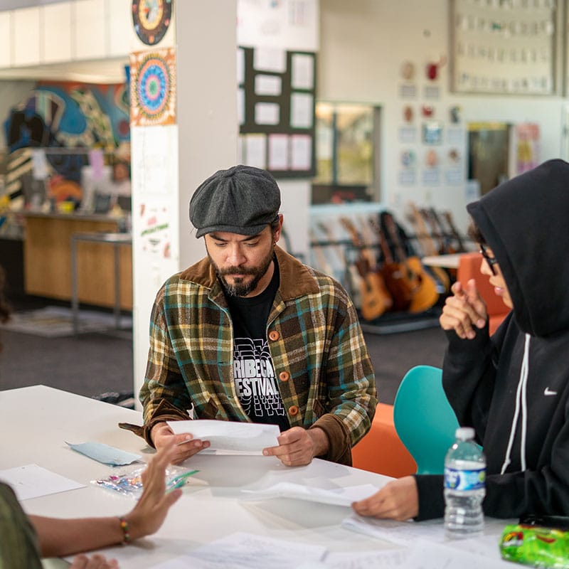 A man in a plaid jacket and flat cap reads papers—perhaps about school attendance—at a table with two others. Guitars, art, and posters decorate the bright, creative room in the background.