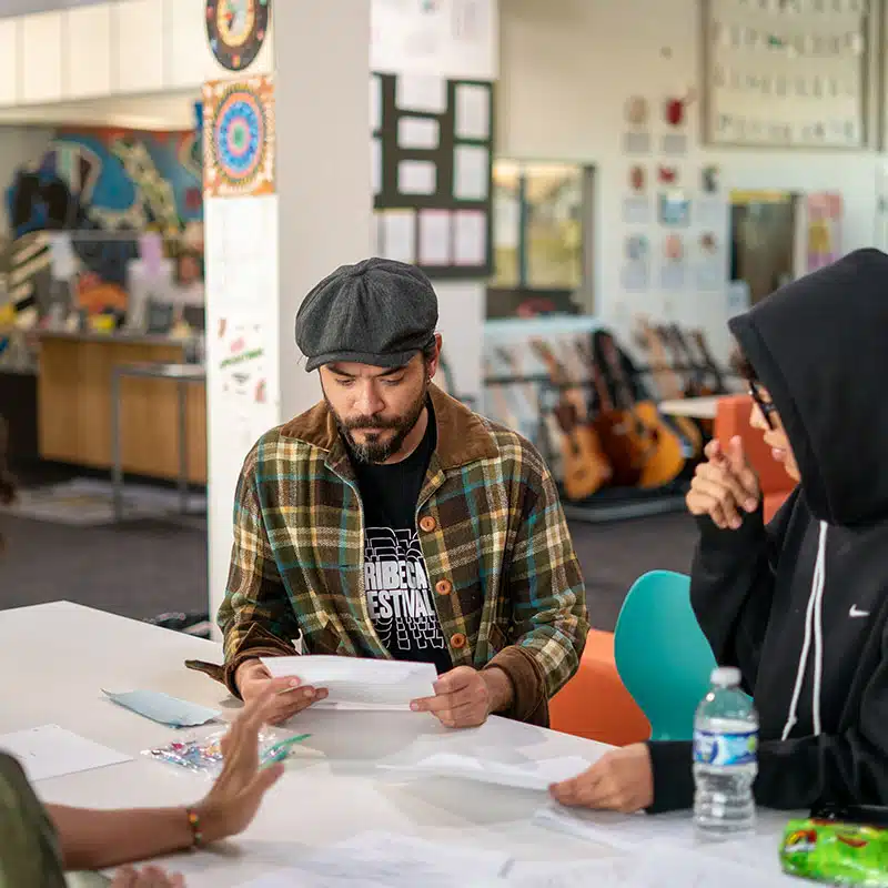 A man in a plaid jacket and flat cap reads papers—perhaps about school attendance—at a table with two others. Guitars, art, and posters decorate the bright, creative room in the background.