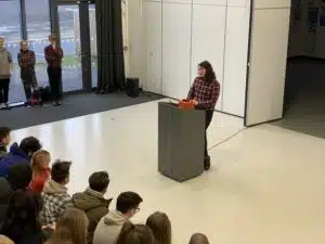 A student in a plaid shirt stands at a podium speaking to a seated audience, emphasizing the importance of student work, while four people stand to the side near a window in a large, bright room.