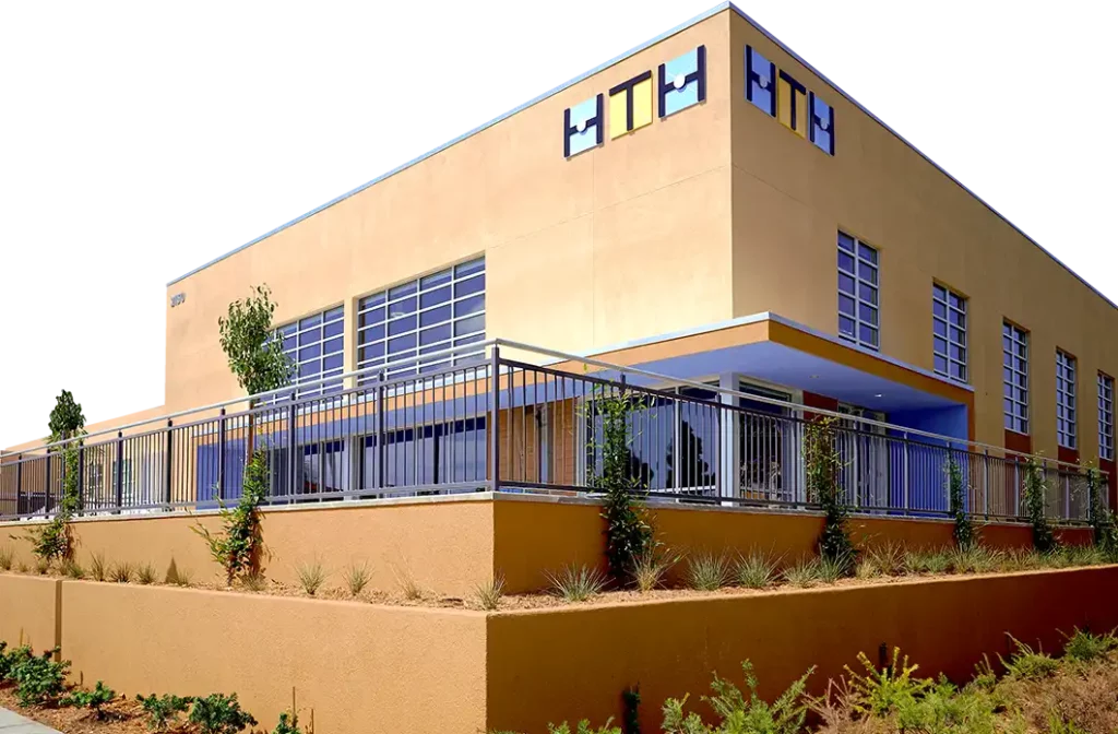 A modern, tan-colored building with large windows, metal railings, and HTH displayed on the upper wall. Desert landscaping surrounds the structure.