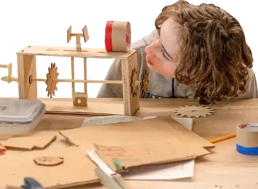 A child with curly hair examines a wooden gear mechanism on a table scattered with papers, gears, a pencil, and craft materials.