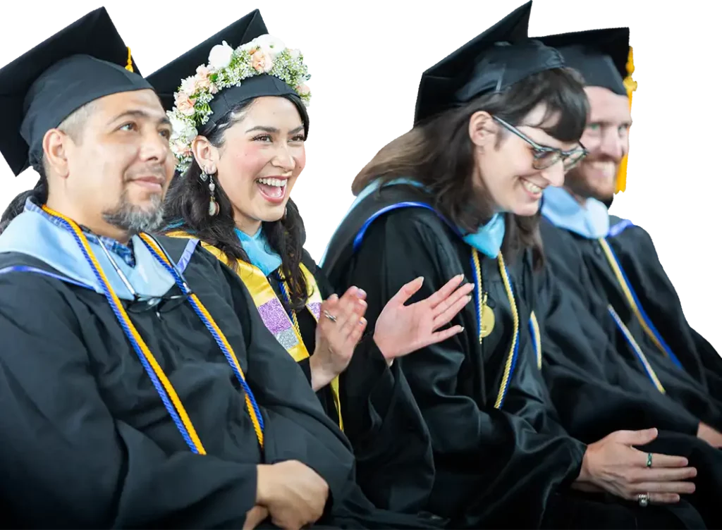 Four graduates in caps and gowns sit in a row. One woman wearing a flower crown is smiling and clapping, while the others sit and smile. The background is white.