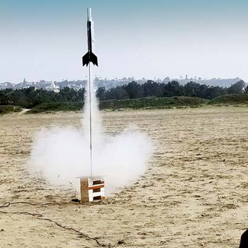 A model rocket, built as part of a project based learning activity, lifts off from a launch pad on a sandy field, trailing a plume of smoke.
