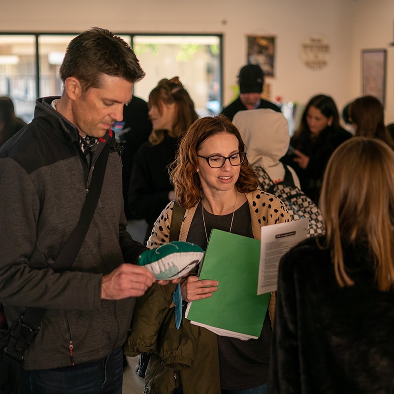 A man and a woman review a document regarding school attendance while holding a stuffed toy fish in a room with others.