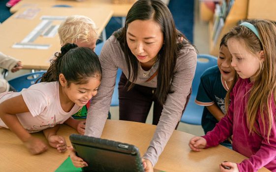 A teacher shows a tablet to a group of young children gathered around a table in a classroom, all of them smiling and exploring the screen together, fostering excitement for continuous improvement in learning. A teacher shows a tablet to a group of young children gathered around a table in a classroom, all of them smiling and exploring the screen together, fostering excitement for continuous improvement in learning.