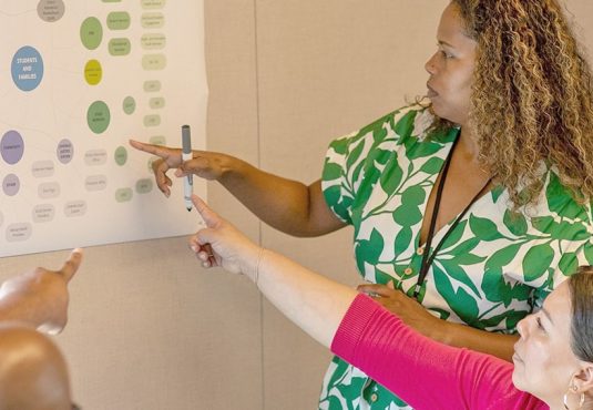 Three people are engaged in a discussion, pointing at a large diagram on the wall. The woman in a green and white patterned dress is explaining how decentralized finance works on the RAISE Network to the seated individuals, who are actively participating.
