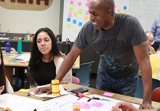 A group of adults collaborates at a classroom table covered with papers and Post-it notes, tackling real problems together. A man stands and smiles, engaging with the group as a teacher talks in the background, fostering continuous improvement.