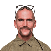 A smiling bald man, identified as Derek Mitchell, with a trimmed mustache and goatee wears glasses on his head, a buttoned olive green shirt, and small earrings. The background is transparent. A smiling bald man, identified as Derek Mitchell, with a trimmed mustache and goatee wears glasses on his head, a buttoned olive green shirt, and small earrings. The background is transparent.