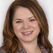 A woman with shoulder-length brown hair smiles at the camera. She is wearing a patterned jacket and a light-colored top, posing against a neutral background for Derek Mitchell’s latest portrait series. A woman with shoulder-length brown hair smiles at the camera. She is wearing a patterned jacket and a light-colored top, posing against a neutral background for Derek Mitchell’s latest portrait series.