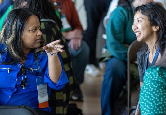 Two women sit facing each other in a crowded room at the Summer Symposium, engaged in an animated conversation. One gestures with her hand while the other smiles, surrounded by other Grade 9 students.